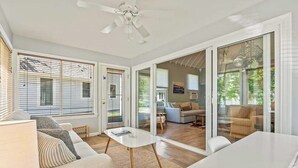 Un vaste espace de vie comprenant un canapé blanc, une table basse et une table à manger. De grandes fenêtres équipées de stores, un ventilateur de plafond et une vue sur la pièce adjacente complètent l’ensemble.