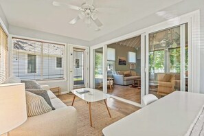 A spacious living area with a white couch, a coffee table, and a dining table. There are large windows with blinds, a ceiling fan, and a view into the adjacent room. - Cozy cottage with steps-to-the-pool access, fully equipped kitchen & beach gear (Wells)