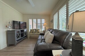 A living room with a leather sofa, a TV on a stand, a lamp, and a window with blinds.