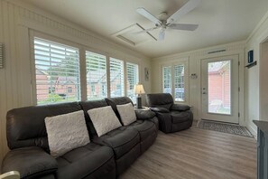 A living room with a dark leather sofa, a ceiling fan, and large windows with blinds.