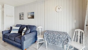 Ein Zimmer mit einem blauen Sofa, einem weißen Sessel, einem Tisch mit gemustertem Tuch und einer Wanduhr.