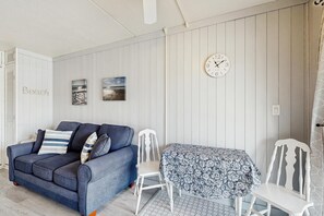 Ein Zimmer mit einem blauen Sofa, einem weißen Sessel, einem Tisch mit gemustertem Tuch und einer Wanduhr.