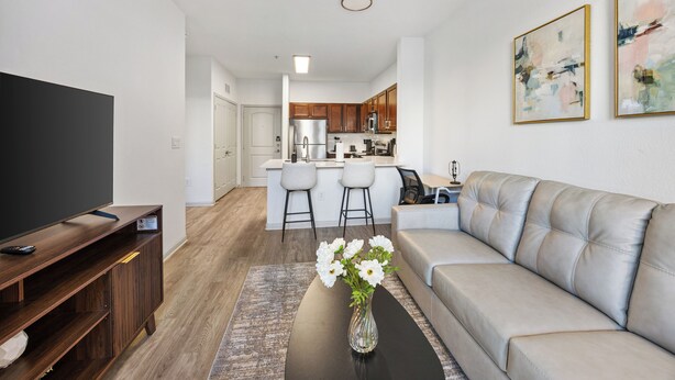 Ein modernes Wohnzimmer mit einer Couch, einem Couchtisch mit Blumen, einer TV-Konsole und einer Küche im Hintergrund.