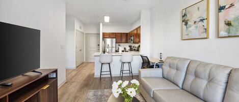 A modern living room with a sofa, a coffee table with flowers, a TV stand, and a kitchen in the background.