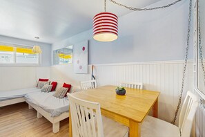 Una habitación con una mesa de comedor de madera, sillas blancas, un banco con cojines, una lámpara colgante y una ventana con persianas amarillas.