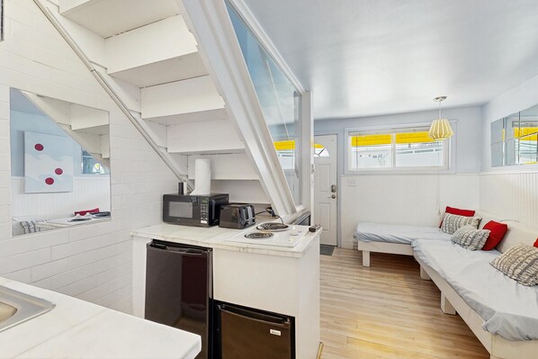 Una habitación compacta con kitchenette, una cama con almohadas, una ventana con persianas amarillas y una escalera.