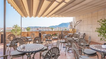 Un patio al aire libre con varias mesas y sillas redondas, un paraguas grande y vistas a edificios y montañas al fondo.