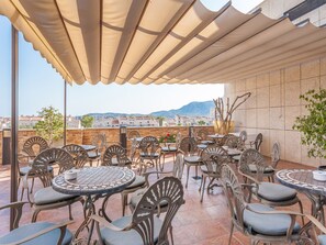 An outdoor patio with multiple round tables and chairs, a large umbrella, and a view of buildings and mountains in the background. - Magic Balneario (Oropesa del Mar)