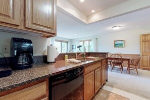 A kitchen with wooden cabinets, a coffee maker, a dishwasher, and a dining area with chairs. - Mountain-view condo with deck,  W/D - close to ski/hike/shopping (Bartlett)