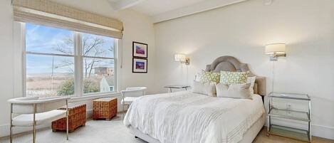 A bedroom with a large bed, a desk, a chair, wicker baskets, and a view of the outdoors.
