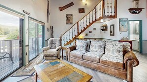Ein geräumiges Wohnzimmer mit einer Holztreppe, einem Sofa mit gemusterten Kissen, einem Fellschaukelstuhl und einem Holztisch.