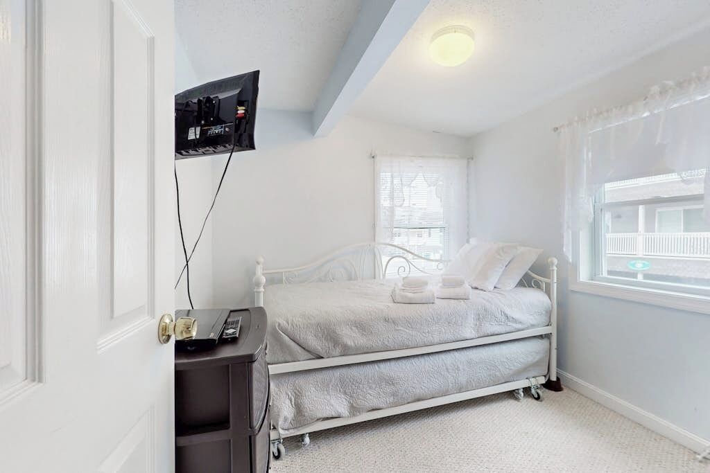Une chambre à coucher avec un lit blanc, un téléviseur fixé au mur, une fenêtre donnant sur un beau paysage et une porte.