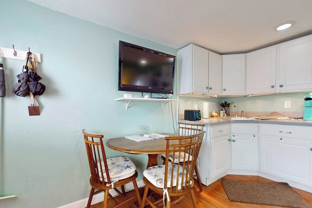 A compact kitchen with white cabinets, a wooden dining table with chairs, a flat-screen TV mounted above the counter, and a small countertop with a kettle and other items.
