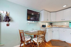 A compact kitchen with white cabinets, a wooden dining table with chairs, a flat-screen TV mounted above the counter, and a small countertop with a kettle and other items. - Cute getaway near the beach with full kitchen, fast WiFi, & furnished deck (Hampton)
