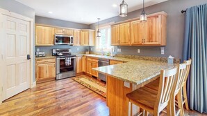 A modern kitchen with wooden cabinets, granite countertops, stainless steel appliances, and a central island with seating. - Chic home with game room, fireplace & mountain view - near Story Land & skiing (Bartlett)