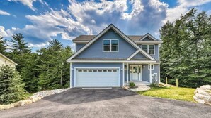 A two-story house with a garage, surrounded by trees and a clear sky. - Chic home with game room, fireplace & mountain view - near Story Land & skiing (Bartlett)