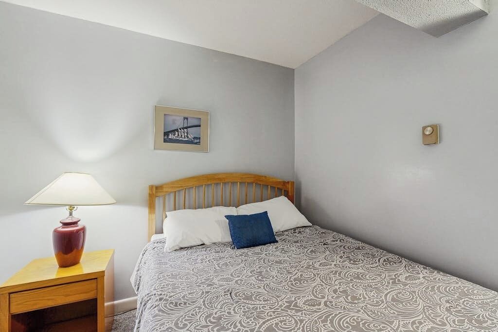 A bedroom with a bed, a nightstand, a lamp, a framed picture on the wall, and a thermostat.