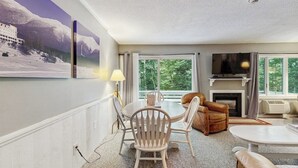 A living room with a fireplace, a television, a dining area with chairs, and a large window with a view of trees.