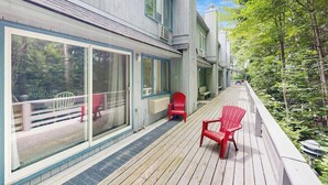 A wooden deck with two red chairs and a bench, a glass door, and a view of a forested area.