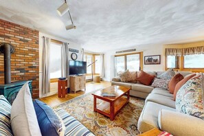 A living room with a brick wall, a fireplace, a sofa, a television, and a large window. - Dog-friendly condo near skiing with amazing Mt.Washington views, fireplace & W/D (Bartlett)