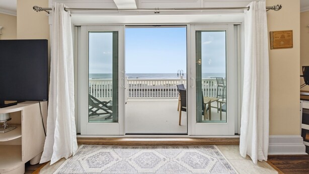 Una habitación con un amplio ventanal corredera que da a un balcón con barandilla blanca, un televisor sobre un mueble bajo y una alfombra estampada en el suelo.