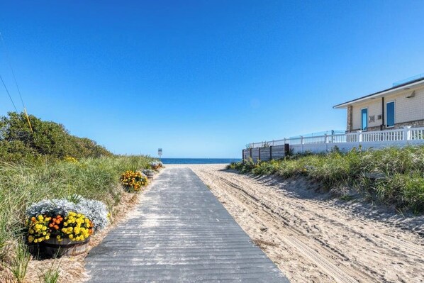A coastal pathway leading to a beach, with a building on the right and a clear sky.