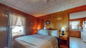 Une chambre à coucher avec un lit, deux lampes de chevet, une fenêtre avec des rideaux, un mur en bois et une vue sur une cuisine.