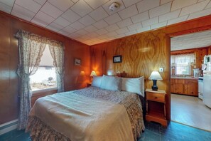 Ein Schlafzimmer mit einem Bett, zwei Nachttischlampen, einem Fenster mit Vorhängen, einer Holzwand und Blick auf eine Küche.