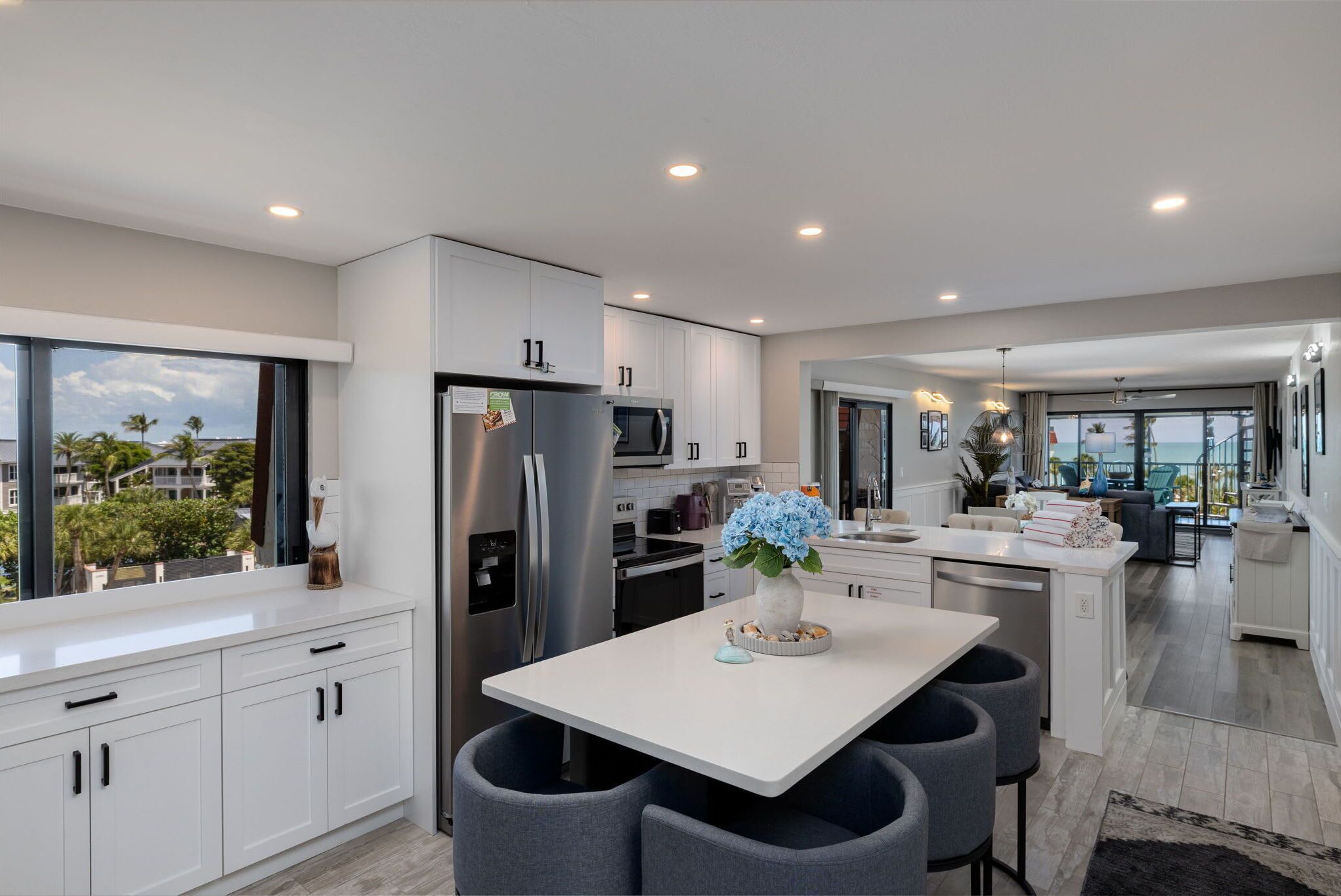 A modern kitchen with white cabinets, a stainless steel refrigerator, a central island with bar stools, and a view of the ocean through large windows.