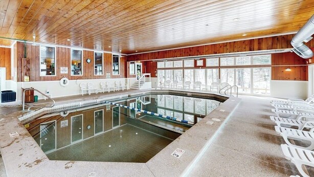 A swimming pool with a wooden ceiling, glass walls, and white chairs along the sides. - Snowbird-friendly home with heated pool, sauna, game room, tennis, BBQ, W/D (Bartlett)