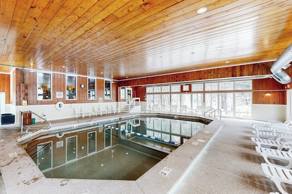 A swimming pool with a wooden ceiling, glass walls, and white chairs along the sides. - Snowbird-friendly home with heated pool, sauna, game room, tennis, BBQ, W/D (Bartlett)