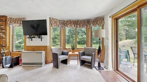 Ein Wohnzimmer mit Kamin, einem Flachbildfernseher, zwei Sesseln, einem Couchtisch und einem großen Fenster mit Blick auf Grünflächen.