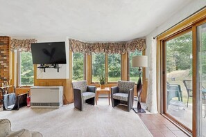 A living room with a fireplace, a flat-screen TV, two armchairs, a coffee table, and a large window with a view of greenery. - Dog-Friendly Condo with Patio, Full Kitchen, W/D, & Free WiFi (Bartlett)