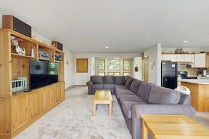 A living room with a sectional sofa, a TV on a wooden cabinet, and a kitchen area with a refrigerator. - Cozy Condo with Shared Pool, High-Speed WiFi, W/D, Partial AC (Bartlett)