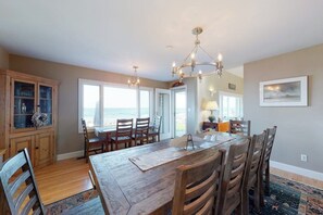 A dining area with a wooden table, chairs, and a chandelier. There is a view of the ocean through large windows. - Oceanfront Stunner w/ Kayaks, Beach Gear, Fireplace, Amazing Water Views & Pond (Rye)