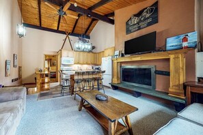 A cozy living room with a fireplace, wooden ceiling beams, a dining area with bar stools, and a kitchen in the background. - Sweet condo with fast WiFi, gas fireplace, & W/D - close to slopes (Bartlett)