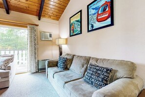 A living room with a sofa, two pillows with alphanumeric codes, a lamp, a window with curtains, and two framed posters on the wall. - Sweet condo with fast WiFi, gas fireplace, & W/D - close to slopes (Bartlett)