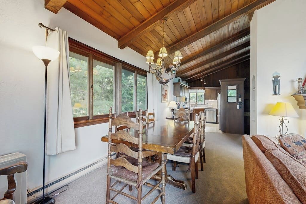 Un comedor con mesa y sillas de madera, una araña y vista a la cocina.