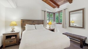 Un dormitorio con una cama grande, cabecera de madera, dos mesitas de noche con lámparas, un banco, una ventana con cortinas y un espejo en la pared.