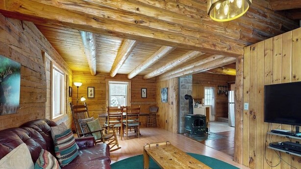 Ein Holzblockhaus-Interieur mit Holzbalkendecke, einem Wohnzimmer mit Sofa und Sesseln, einem Essbereich und einer Küche mit Herd.