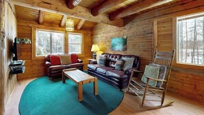 Ein rustikales Blockhaus-Interieur mit Holzbalkendecke, zwei Sofas, einem Schaukelstuhl und einem großen Fenster.