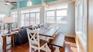 Un comedor con una mesa de madera, sillas blancas, un sofá y ventanas amplias.