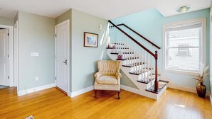Una habitación con piso de madera, una escalera de madera, un sillón color beige y un cuadro en la pared.