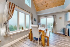 Ein Essbereich mit Holztisch und -stühlen, großen Fenstern mit Vorhängen und Blick ins Freie.