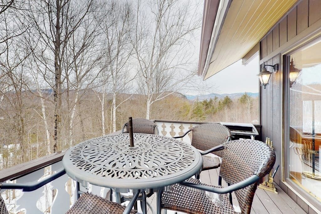 Een overdekt terras met een ronde tafel en rieten stoelen, met uitzicht op een landschap met bomen en heuvels in de verte.