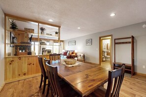Ein Essbereich mit Holztisch und -stühlen, eine Küche mit Holzschränken sowie ein Wohnzimmer mit einer Couch und einem Fernseher.