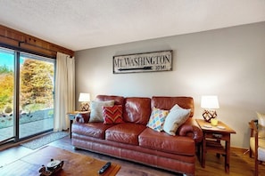 Ein Wohnzimmer mit einem braunen Ledersofa, zwei Beistelltischen mit Lampen, einem Schild mit der Aufschrift 'MT WASHINGTON 10 MI.' und einem großen Fenster mit Blick auf Bäume.