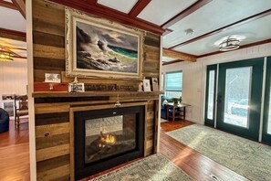 Un salon avec une cheminée, des boiseries, un tableau encadré et une vue à travers une porte vitrée.