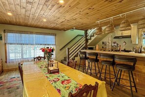 Un espace repas comprenant une longue table, des tabourets de bar, un escalier et un coin cuisine.