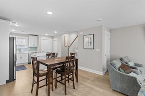 A modern kitchen with white cabinets, a dining table with chairs, a gray sofa, and a staircase.